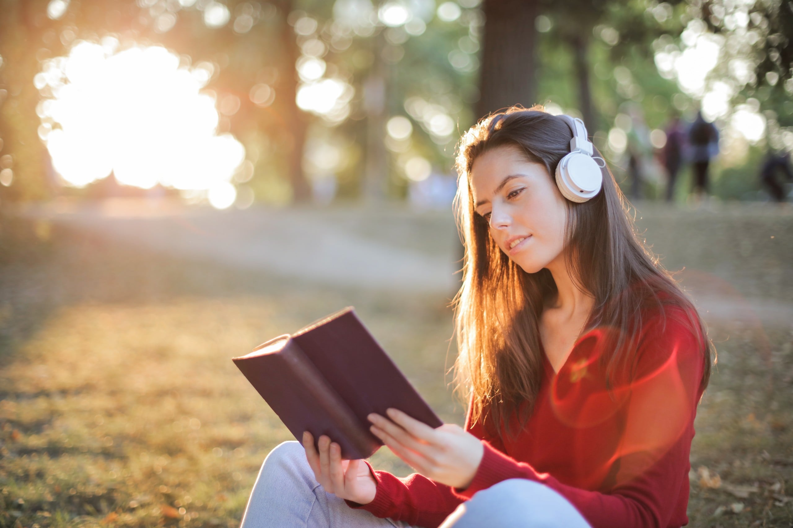 A woman enjoying nature, wearing headphones, and engrossed in a book while sitting on the grass.